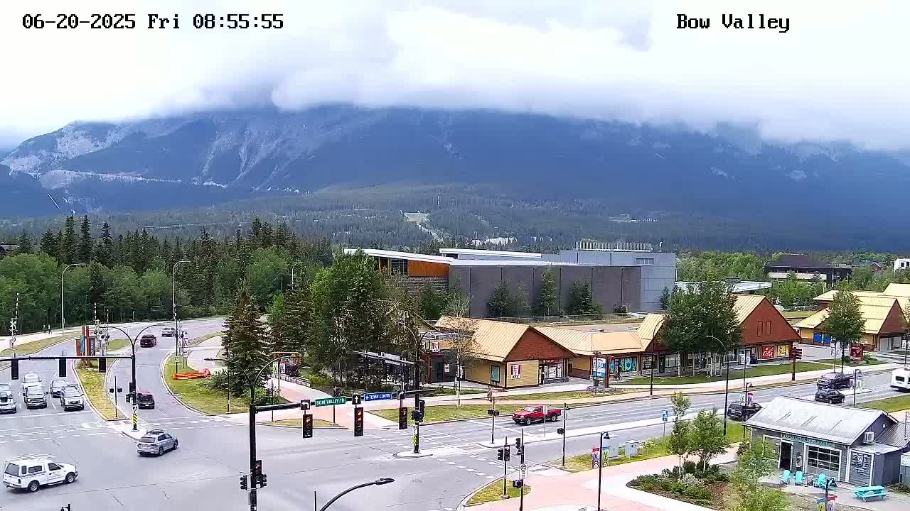 A partly cloudy day reveals a mountain range in the background, overlooking a roadway intersection with several cars and buildings, including some small shops.