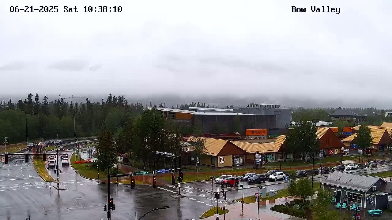 A wet, overcast day reveals a view of a town's intersection, with several buildings and vehicles visible, and a line of trees and distant mountains obscured by fog in the background.