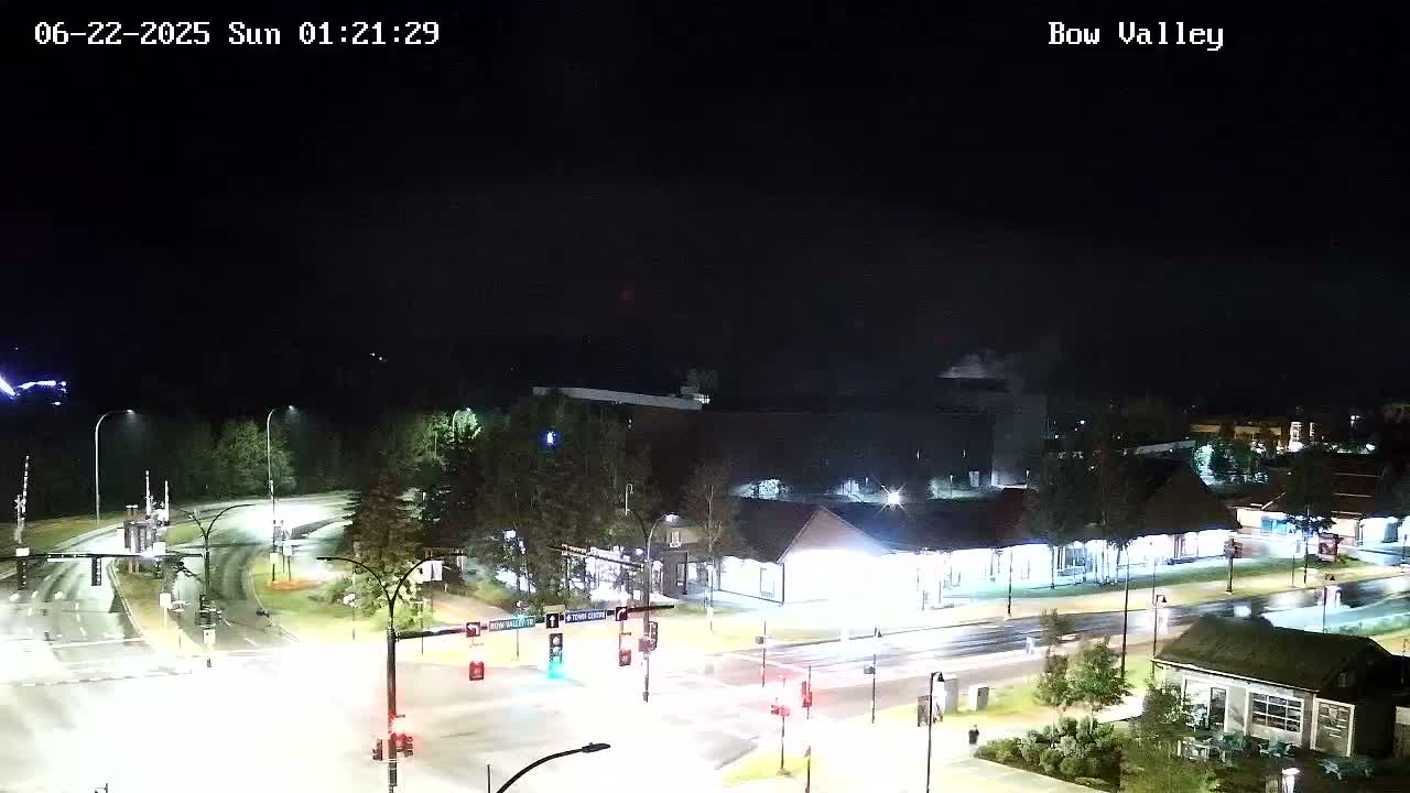 A wet, nighttime view of a road intersection and surrounding buildings, with light rain falling.