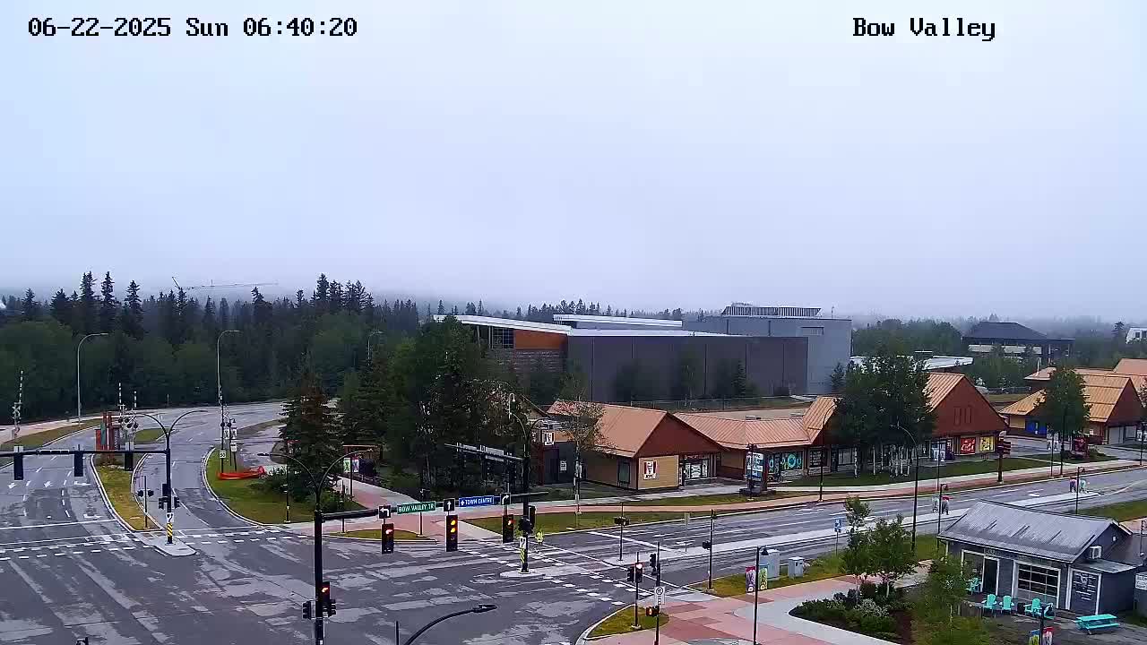 A hazy, overcast view shows a road intersection with several buildings, including a large gray structure and smaller, reddish-brown commercial buildings, surrounded by green trees.