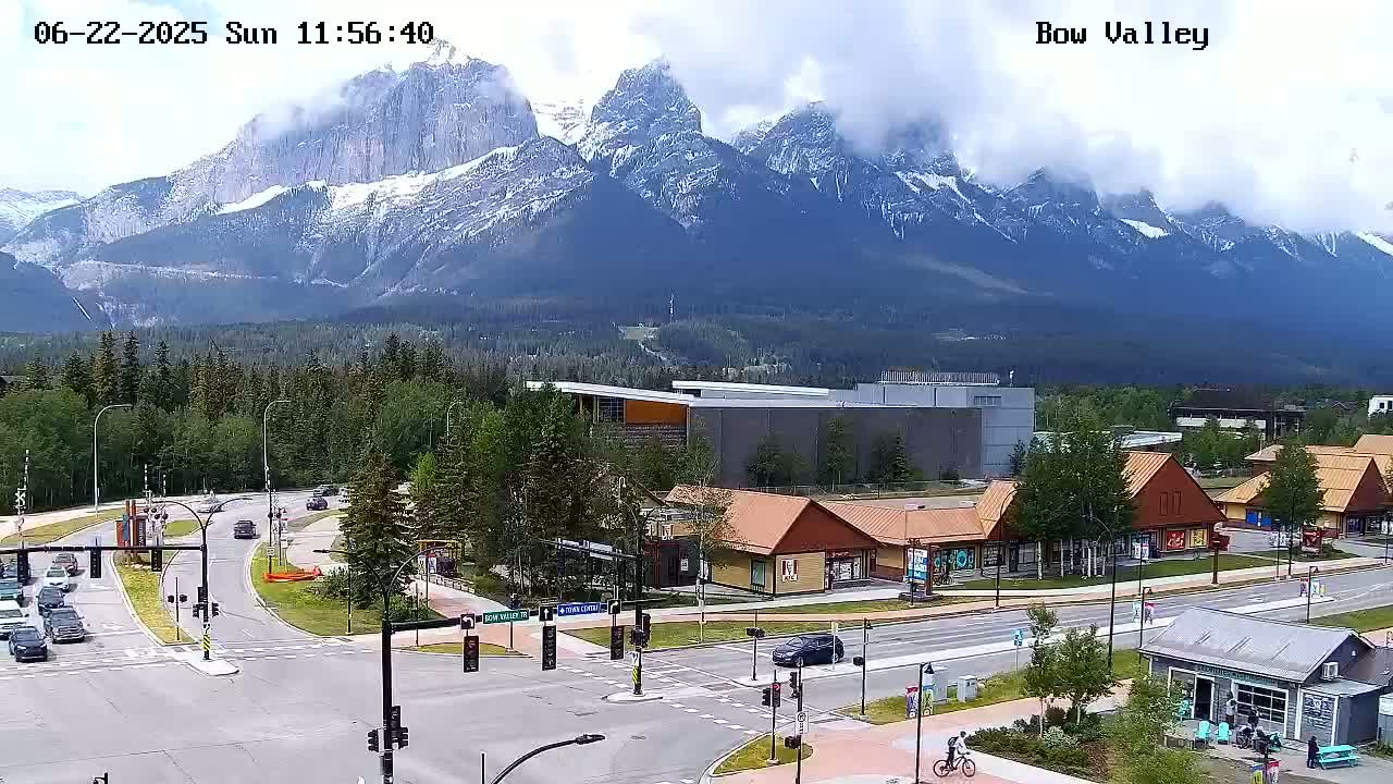 A partly cloudy day reveals snow-capped mountains in the background of a town with a road intersection and several buildings, including shops and a larger building.
