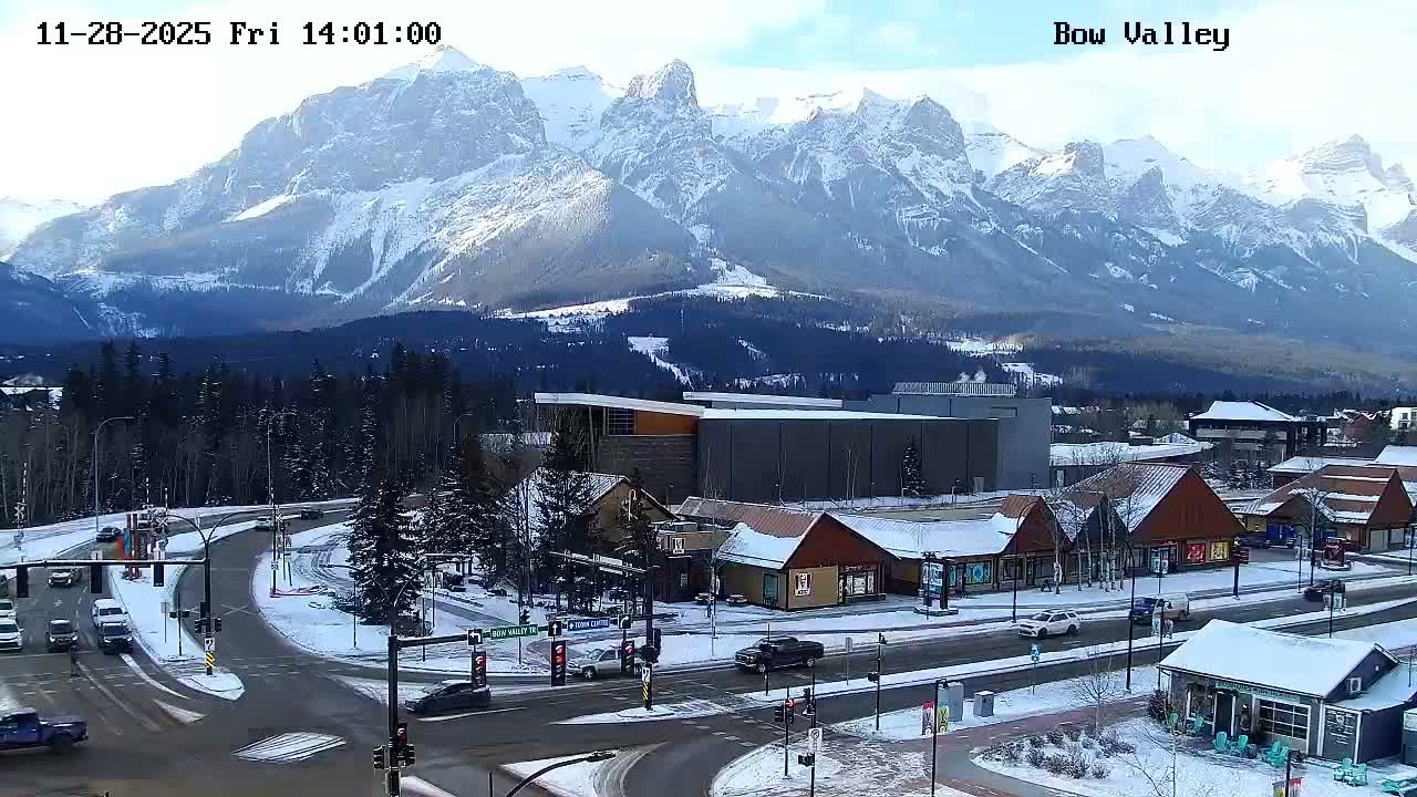 Majestic snow-capped mountains loom over a picturesque snow-covered town with roads and vehicles, all under a bright, clear winter sky.