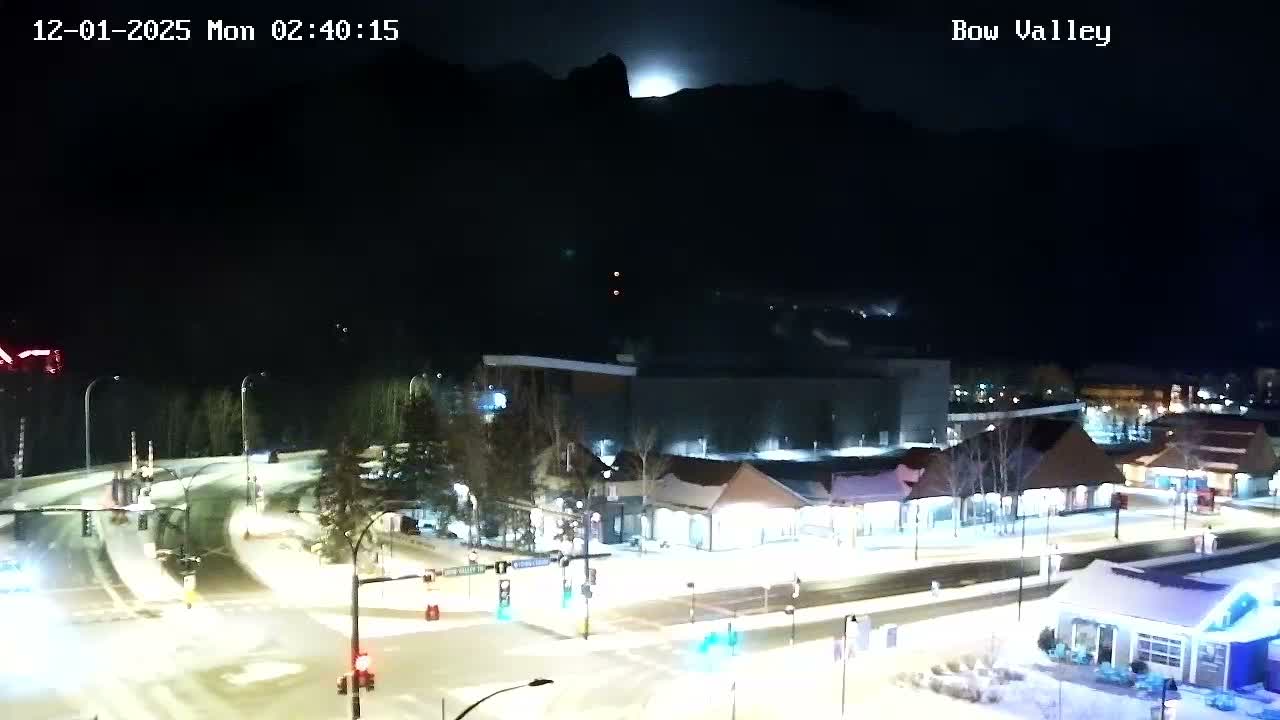 A snow-covered town is seen at night from an elevated perspective, illuminated by streetlights and building lights, with dark mountains silhouetted against a clear sky where a bright full moon is visible above a peak.
