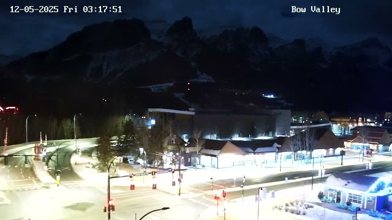 The image shows a snowy mountain town at night under clear, cold conditions, with illuminated roads and buildings set against a backdrop of dark, rugged peaks.