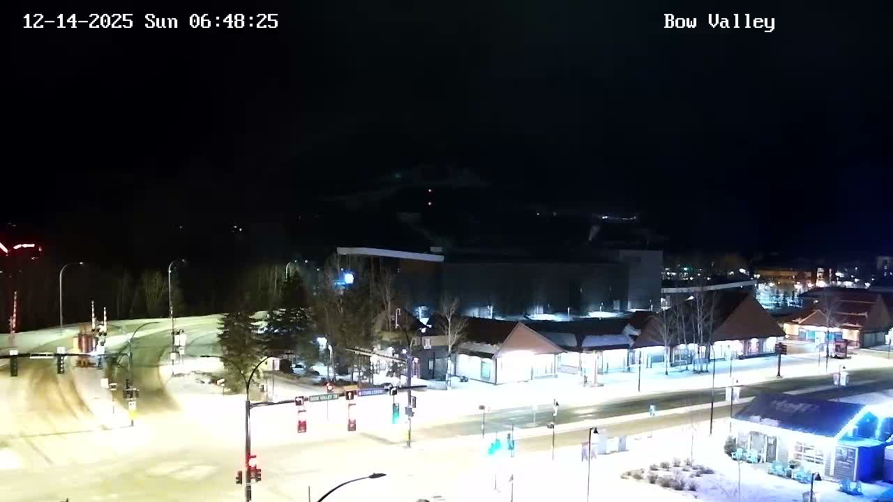 The image shows a snowy mountain town at night under clear, cold conditions, with illuminated roads and buildings set against a backdrop of dark, rugged peaks.