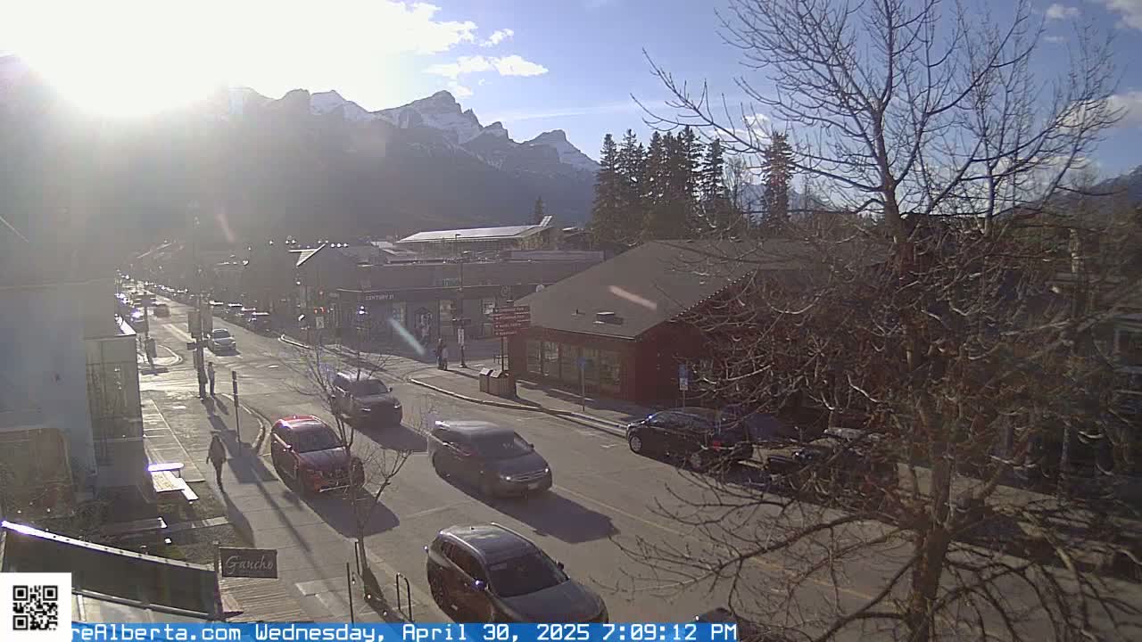 A sunny day in a mountain town shows a street scene with several cars and pedestrians, under a bright sky with snow-capped mountains in the background.