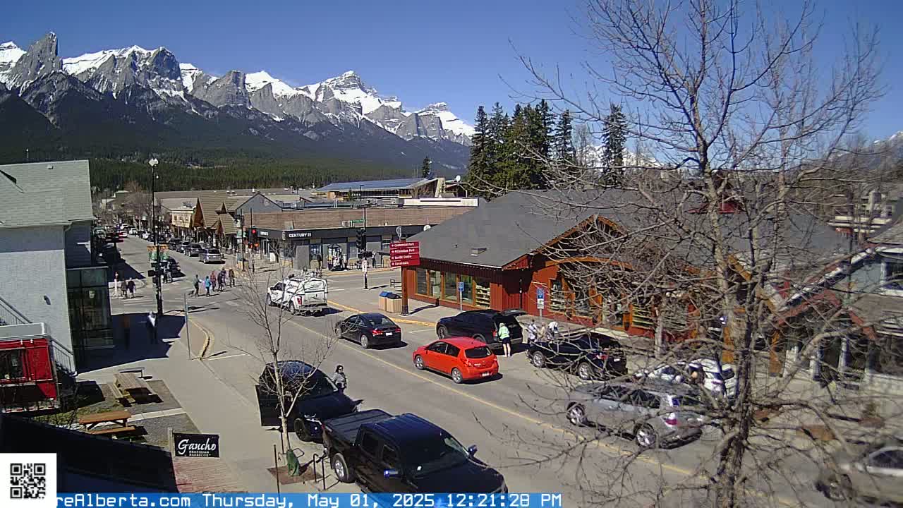 A sunny day shows a mountain view overlooking a town's street with cars parked and pedestrians walking, along with several shops and buildings.