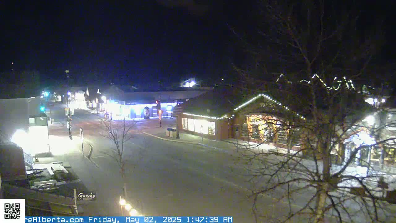 A nighttime, snow-covered street scene shows several illuminated buildings with strings of lights, bare trees, and a mostly empty roadway.