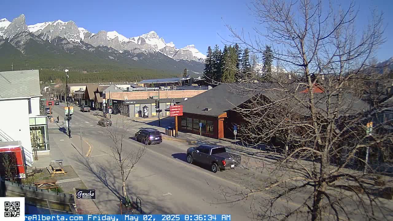 A sunny day reveals a mountain range in the background, with a town's street scene in the foreground, showing vehicles on a road lined with buildings and bare trees.