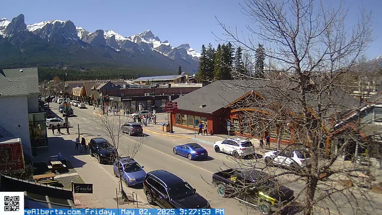 A sunny mountain town street scene with cars parked and driving along a road lined with shops and buildings, under a clear sky and snow-capped mountains in the background.