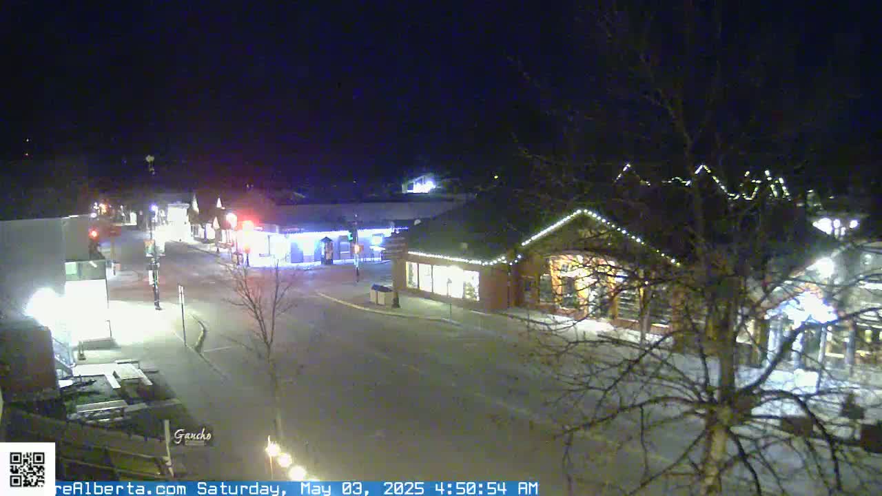 A nighttime street scene shows a quiet town square with several illuminated buildings, bare trees, and a few cars.