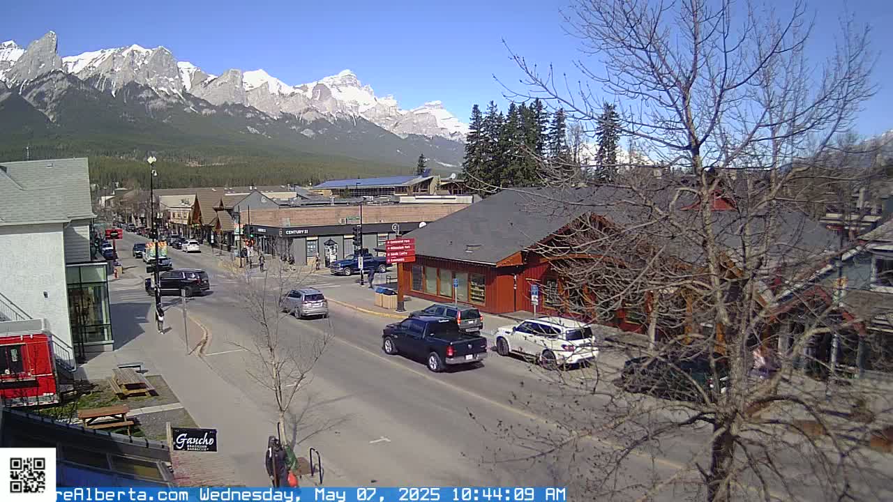 A sunny day shows a mountain view from a street in a town with cars driving on the road and pedestrians walking on the sidewalk, bare trees lining the streets.