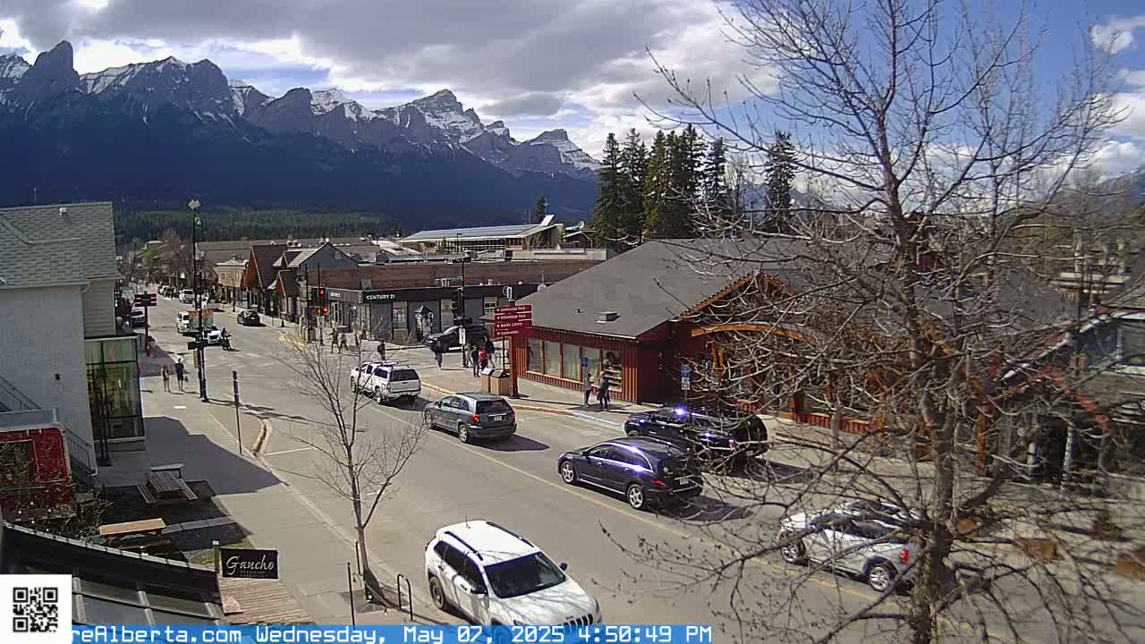 A partly cloudy day reveals a mountain range in the background of a town's main street, where cars are parked and driving on the road, with people walking on the sidewalk.