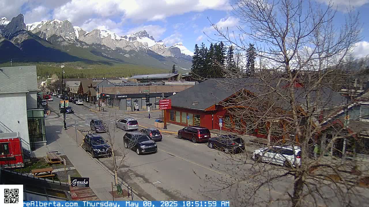 A partly cloudy day reveals a mountain range behind a town's main street where cars are driving along the road.