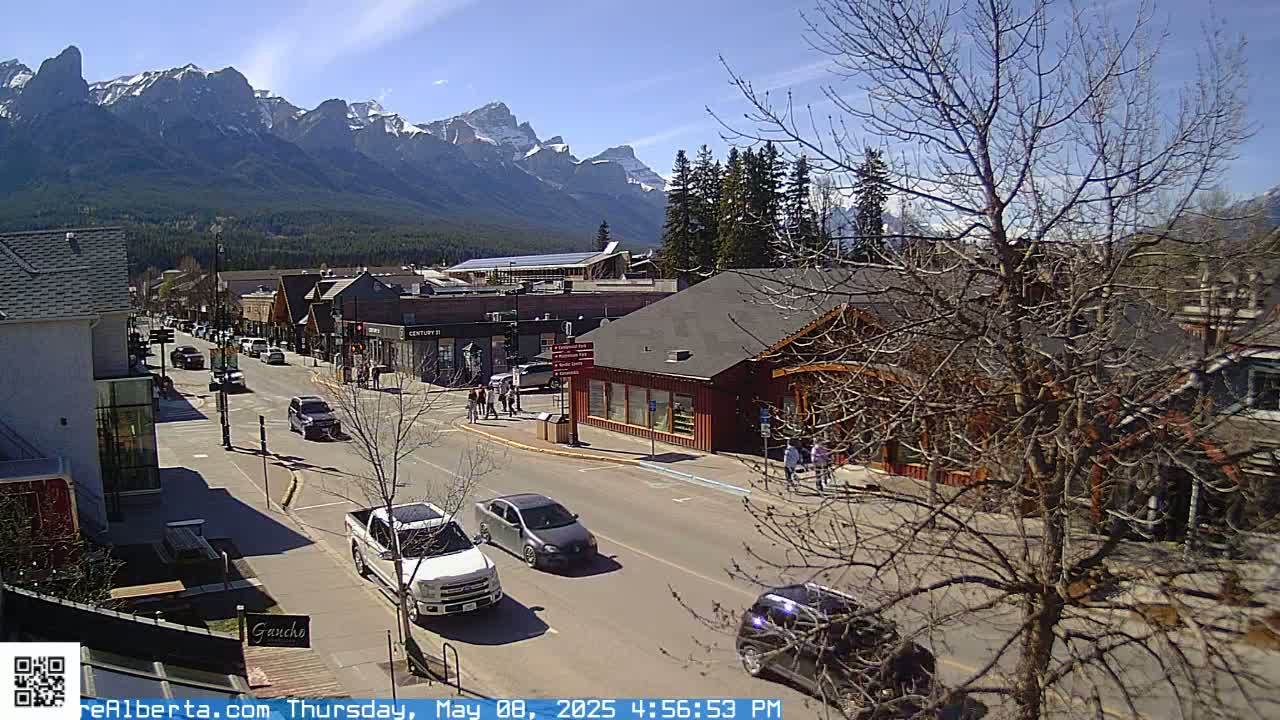 A sunny day reveals a mountain vista overlooking a town's street scene with cars parked and driving along the road, in front of buildings with pedestrians walking.
