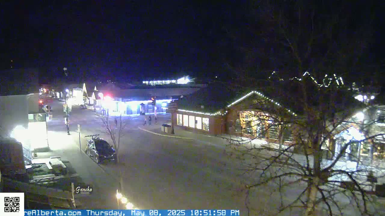 A nighttime, street-level view of a town square with several illuminated buildings, a few cars parked, and bare trees.