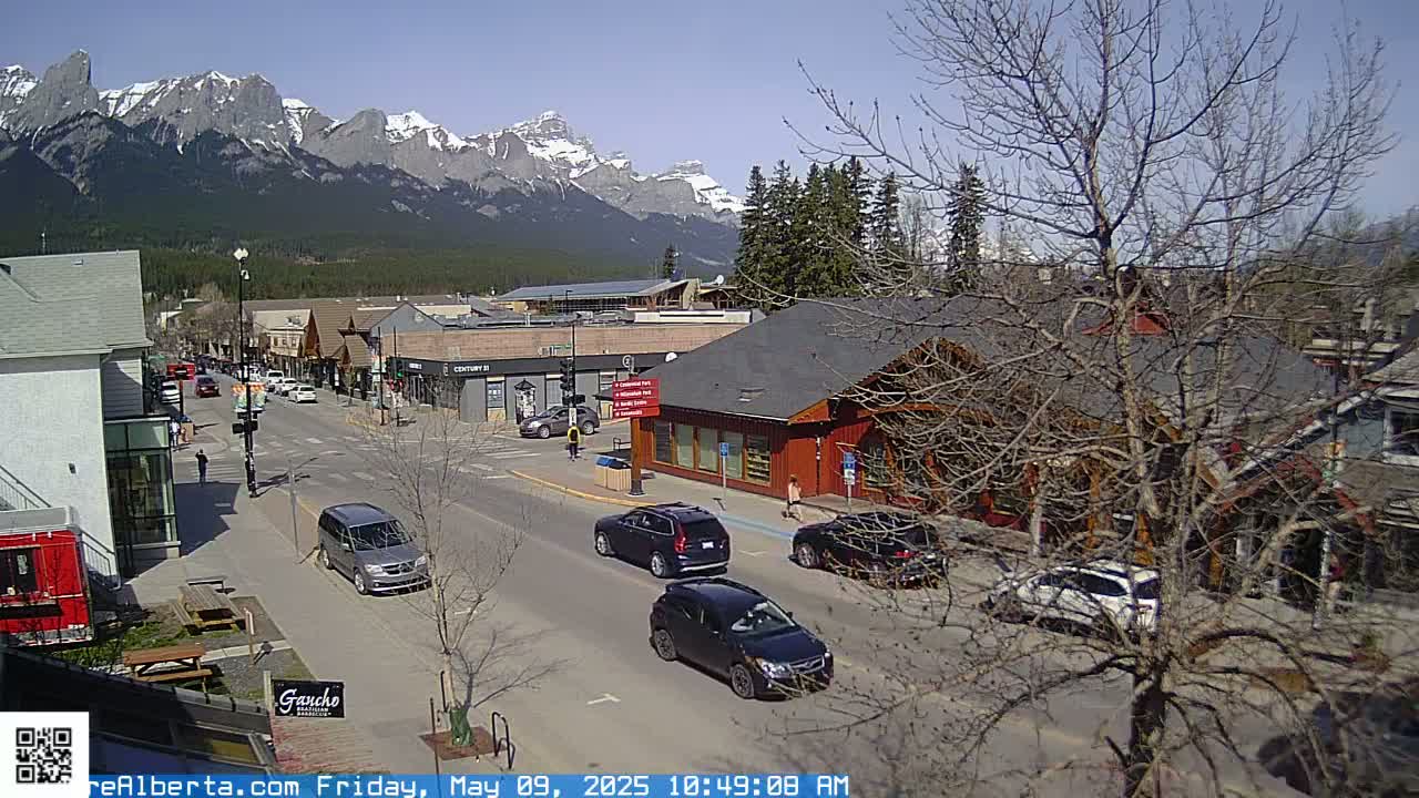 A sunny day reveals a mountain range in the background, overlooking a town's main street with several cars parked and driving along it.
