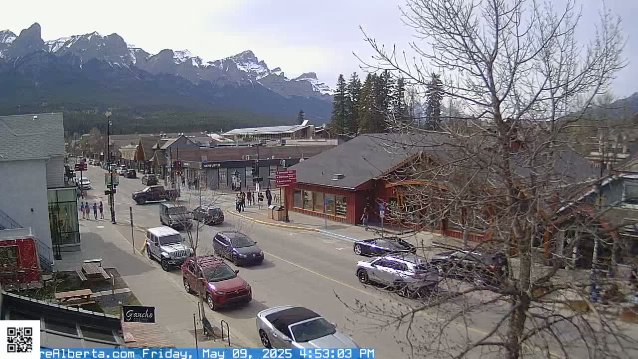A street scene shows several cars parked and driving on a road lined with shops and buildings, set against a backdrop of mountains on a partly cloudy day.