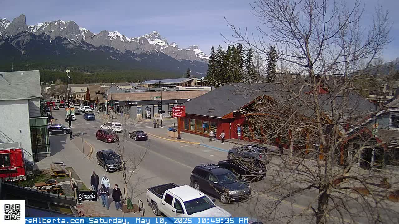 A sunny day shows a mountain view overlooking a town's street with cars parked and driving, and pedestrians walking.