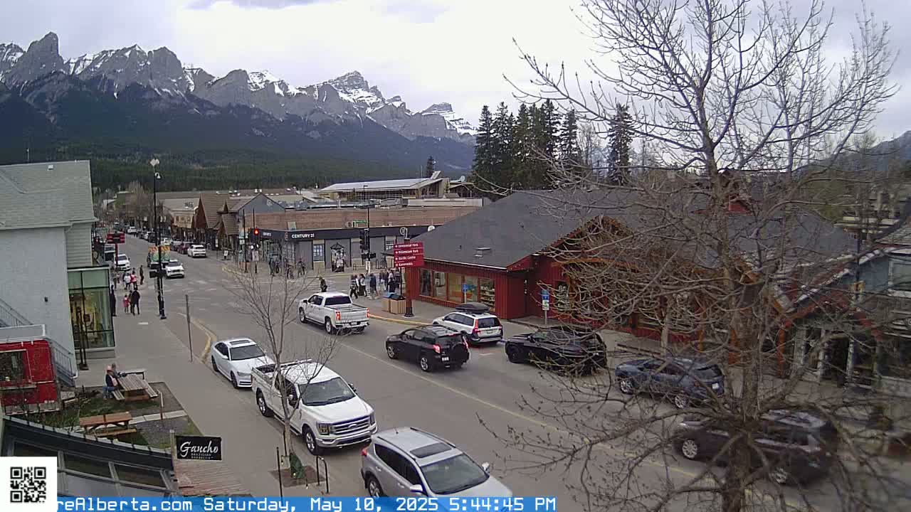 A mountain town street scene with overcast skies shows cars parked and driving along a road lined with shops and buildings, with snow-capped mountains visible in the background.
