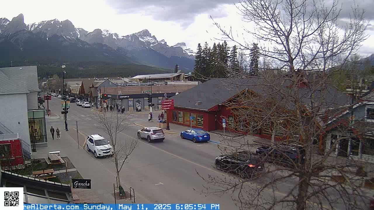 A street scene in a town nestled against a mountain range under an overcast sky shows several cars on a street lined with buildings and pedestrians walking along the sidewalk.