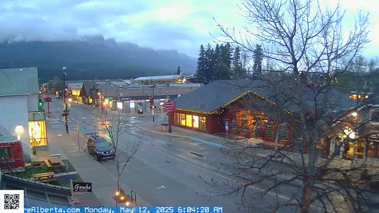 A wet street scene in a mountain town at dawn shows several buildings, cars, and bare trees under an overcast sky with mountains in the background.