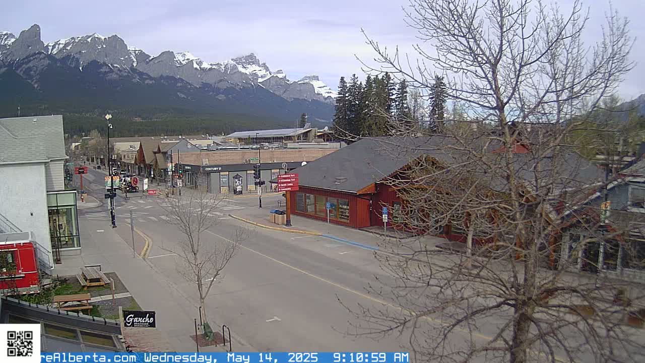 A mostly empty street in a town nestled in a valley, with snow-capped mountains in the background under a cloudy sky.