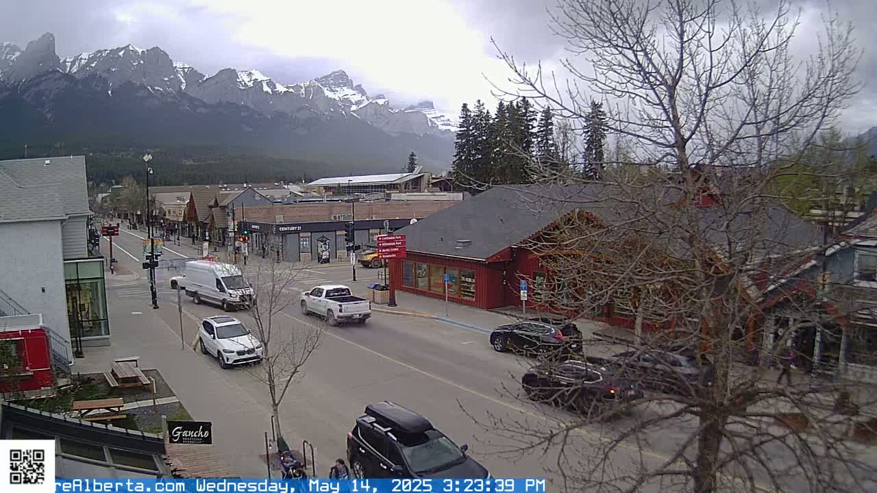 A street scene shows several vehicles driving on a road lined with shops and buildings, with snow-capped mountains in the background under an overcast sky.
