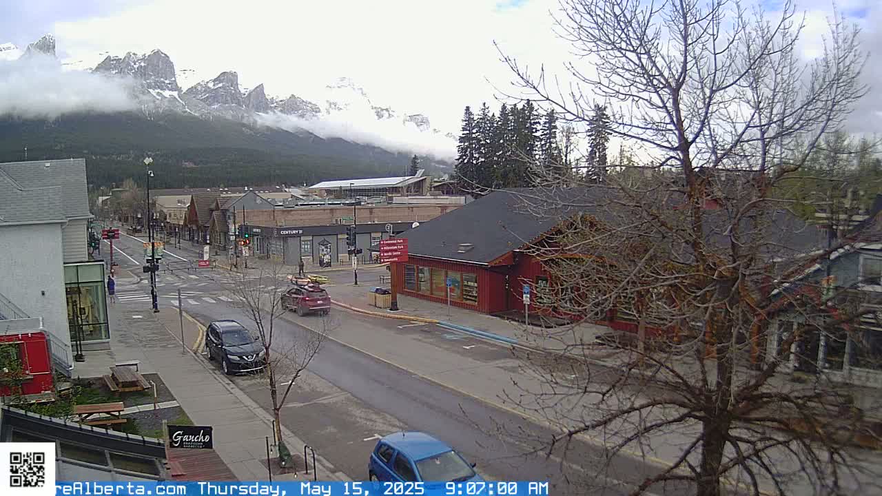 A partly cloudy day reveals a mountain range shrouded in mist behind a town's street scene with several cars and bare trees.