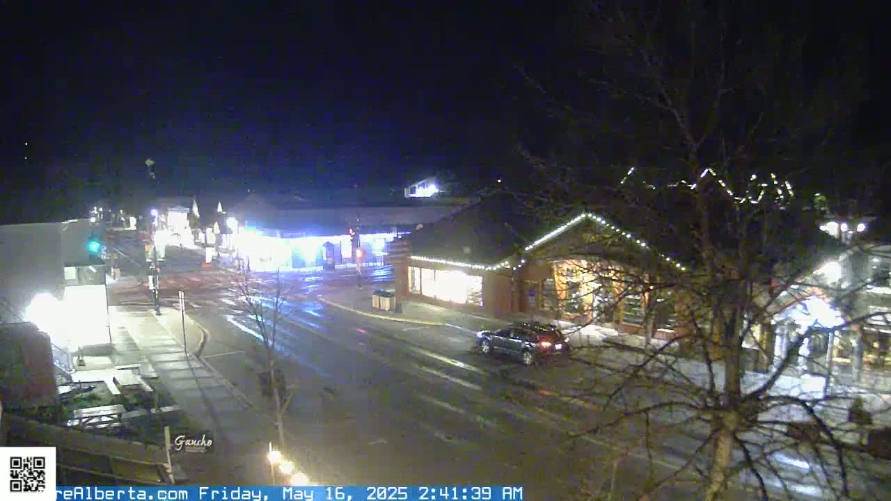 A nighttime view of a town square with wet roads, showing a vehicle driving down the street past buildings with lights on.