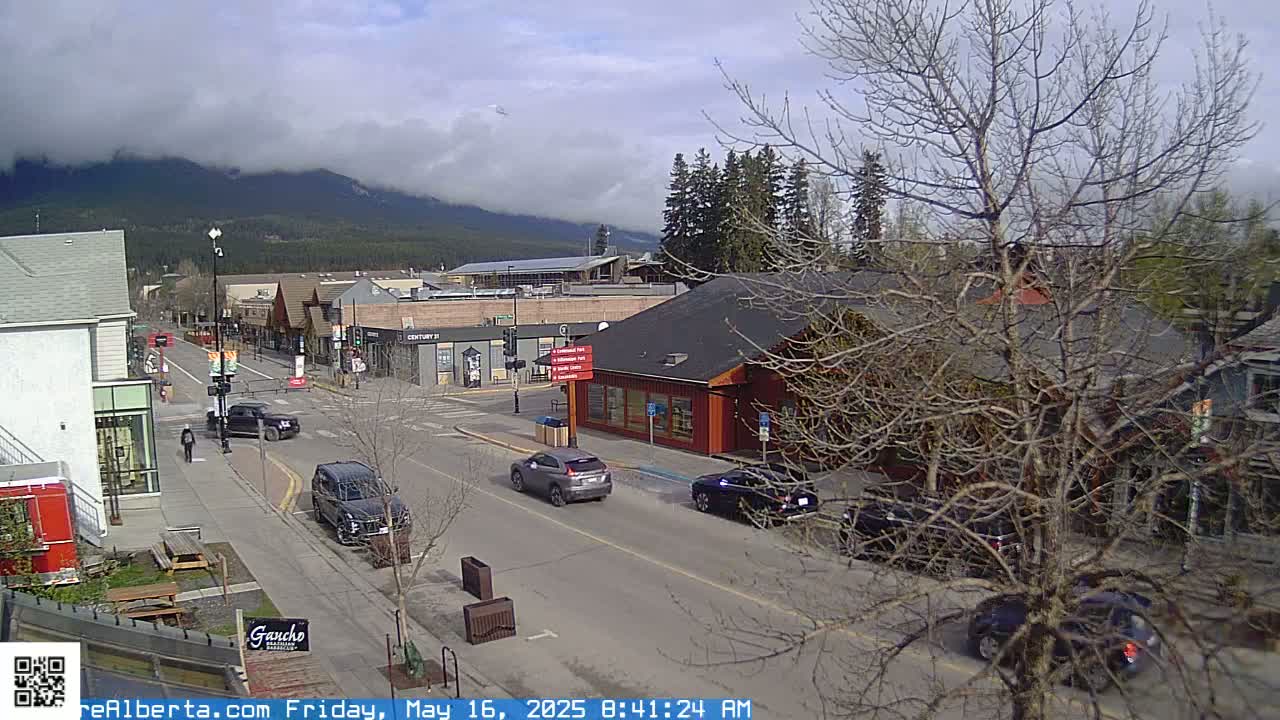 A partly cloudy day reveals a mountain range in the background of a town's street scene with cars parked and driving along a road lined with shops and buildings.