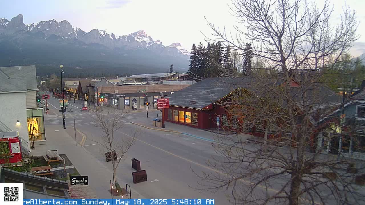 A mostly empty street in a town at the base of snow-capped mountains on a cloudy morning.