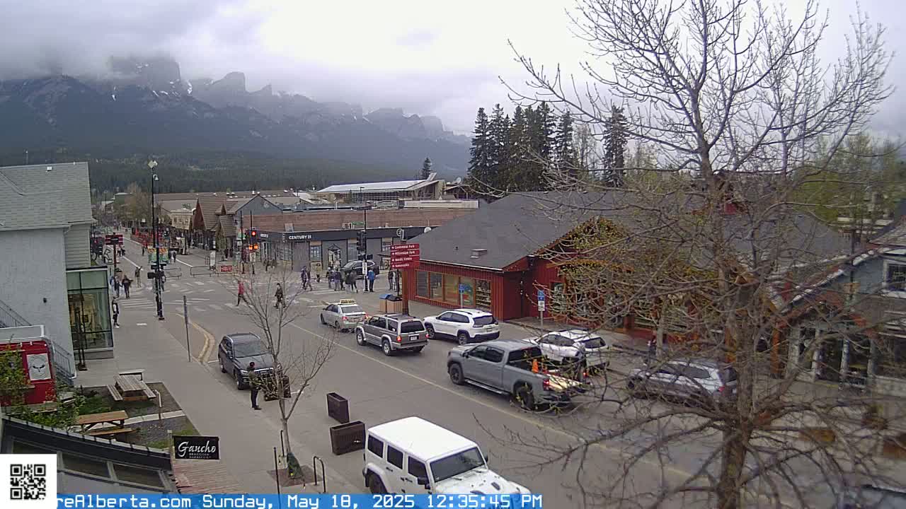 A street scene in a mountain town on an overcast day shows cars driving on a road lined with shops and buildings, with pedestrians walking on the sidewalks and mountains in the background.