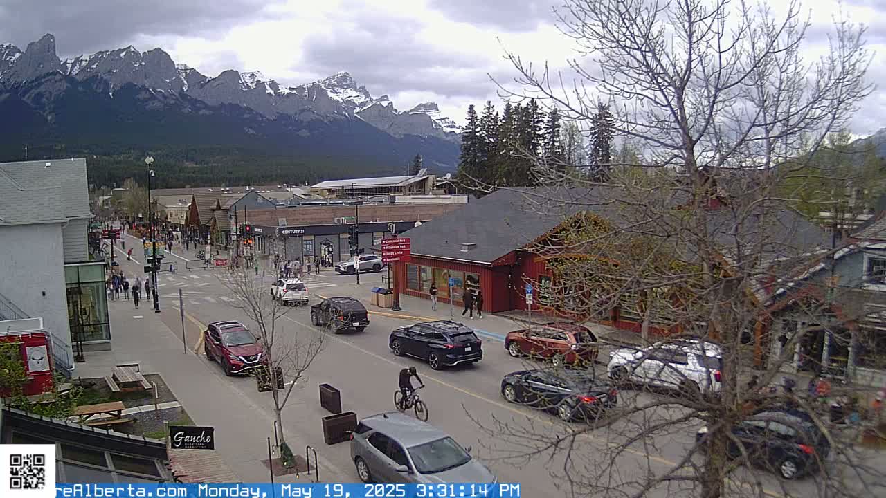 A street scene in a mountain town shows several cars and pedestrians on a road, with snow-capped mountains in the background under a cloudy sky.