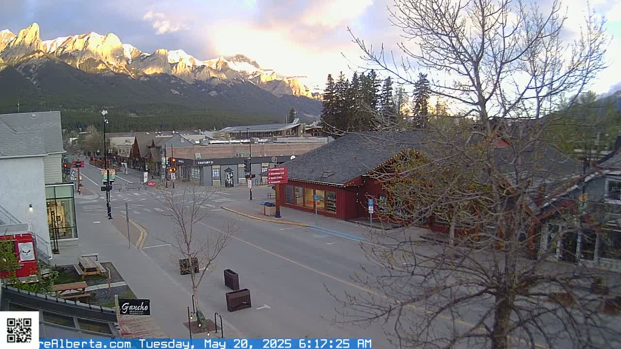 A mostly empty street in a mountain town at dawn, with snow-capped mountains in the background under a partly cloudy sky.