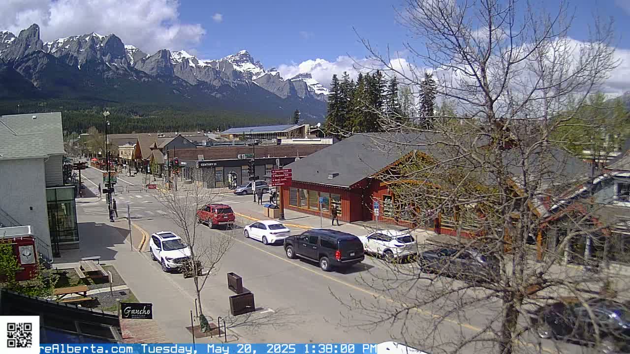 A partly cloudy day reveals a mountain range in the background of a town's street scene with several vehicles parked and driving on the road.