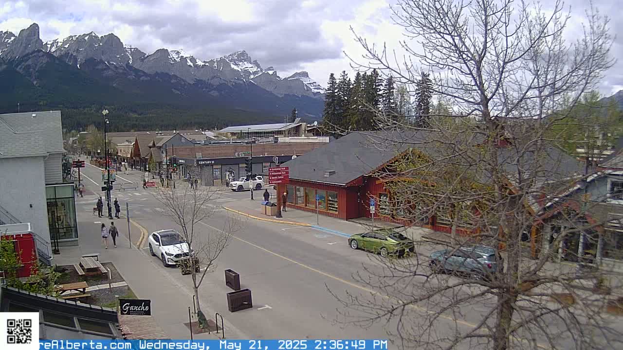 A partly cloudy day shows a mountain view overlooking a town's street with cars and pedestrians, some trees are bare.