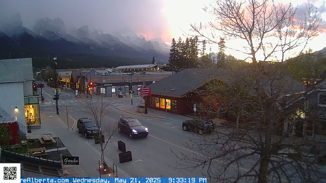 A mountain town street scene at sunset shows several cars driving past shops and restaurants, with mountains in the background under a partly cloudy sky.