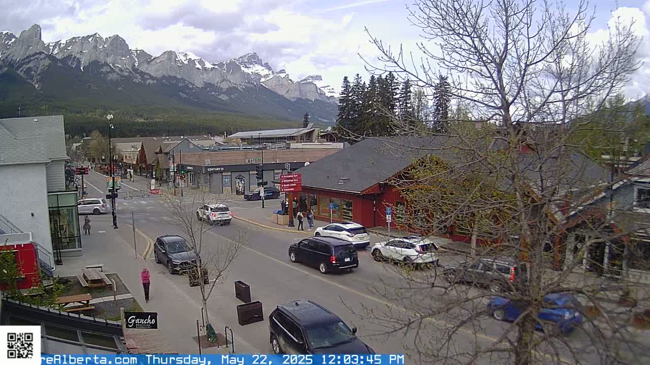 A partly cloudy day reveals a mountain range in the background of a town's street scene, where several cars are parked and driving along a road lined with shops and buildings.