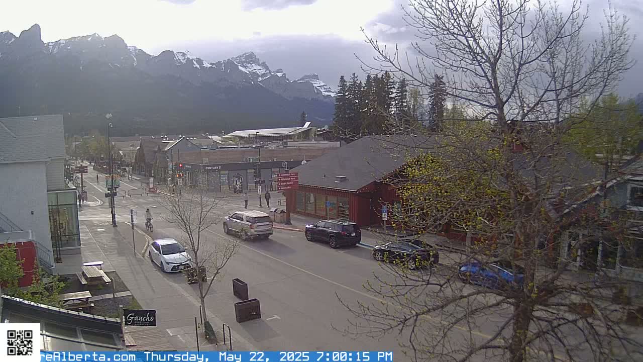 A street scene in a town nestled in a valley with snow-capped mountains in the background under an overcast sky.