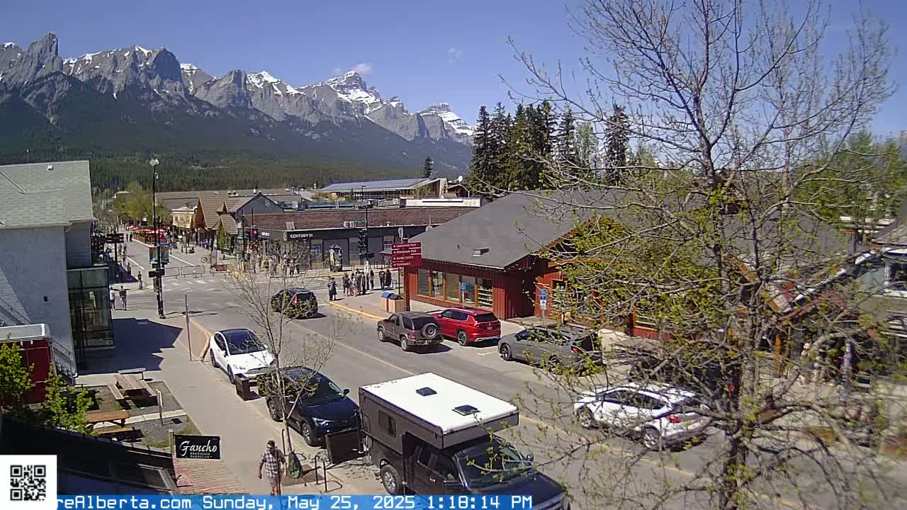 A sunny day reveals a mountain view overlooking a town's main street, where cars are parked and pedestrians stroll amidst shops and buildings.