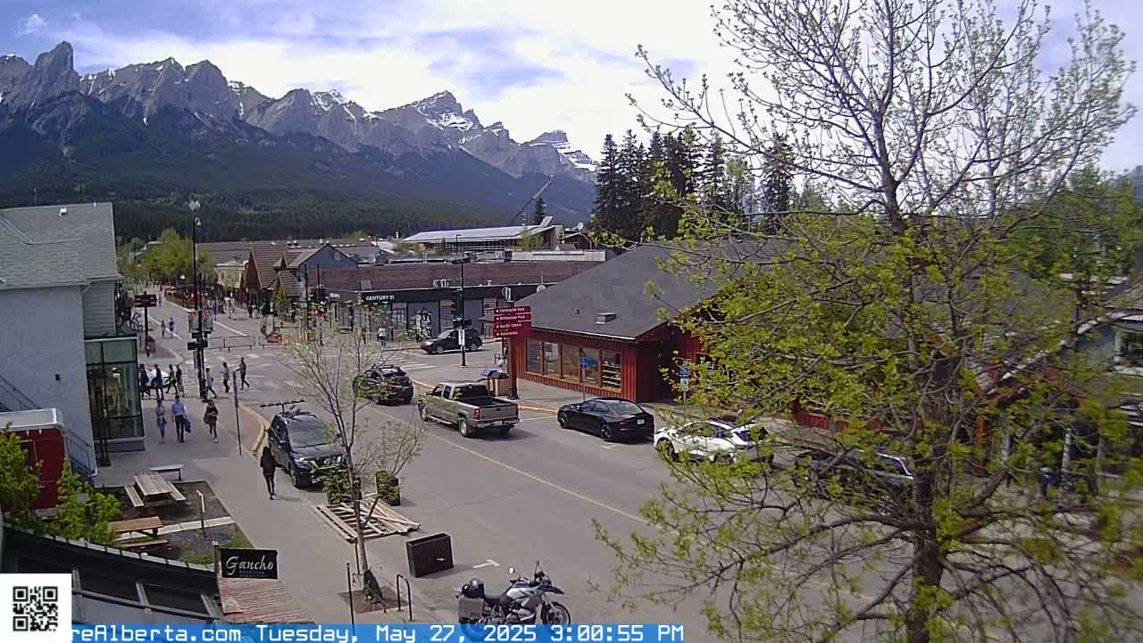 A street scene in a town nestled against a mountain range under a partly sunny sky shows pedestrians walking and cars driving on a street lined with buildings.