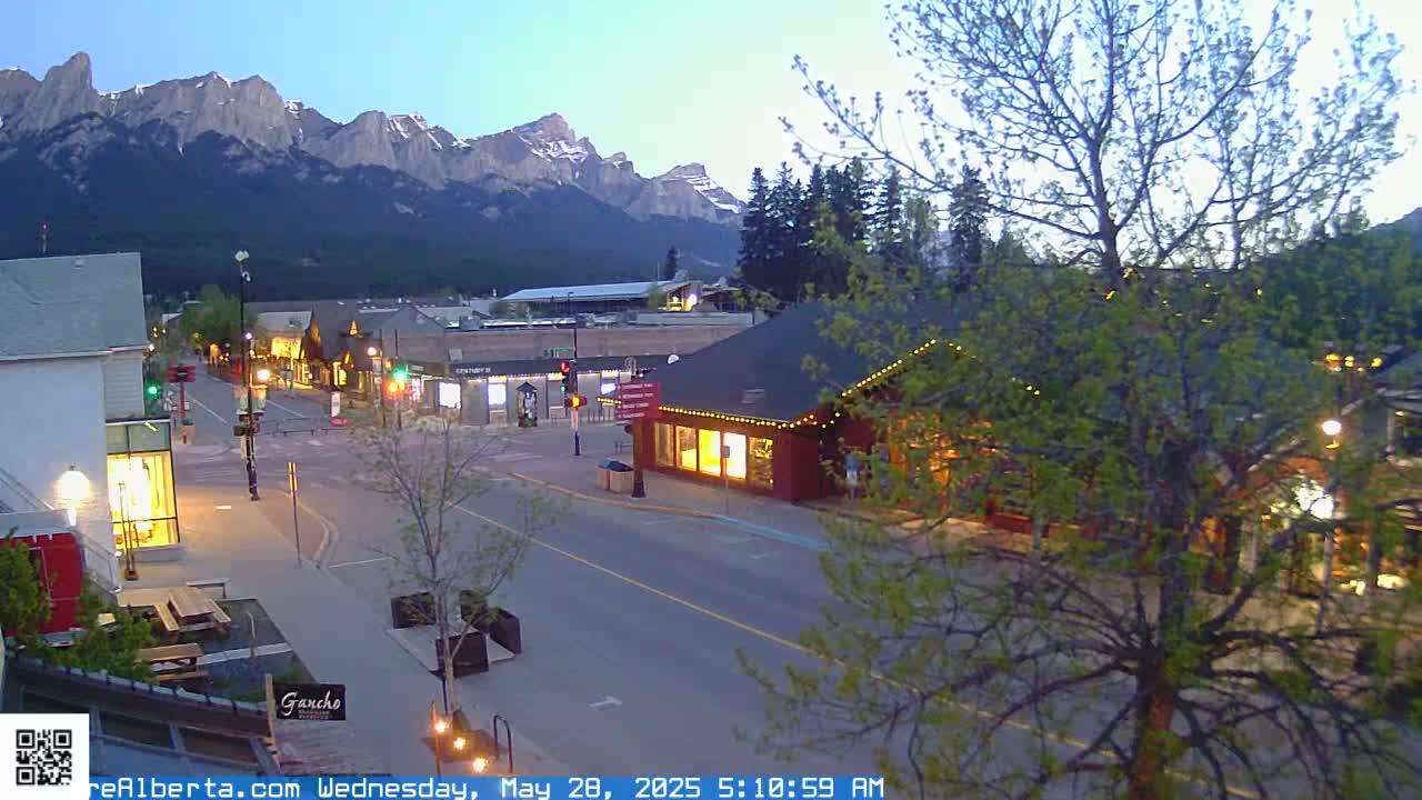 A twilight view of a town's main street, with buildings lit up, under a clear sky, and mountains in the background.