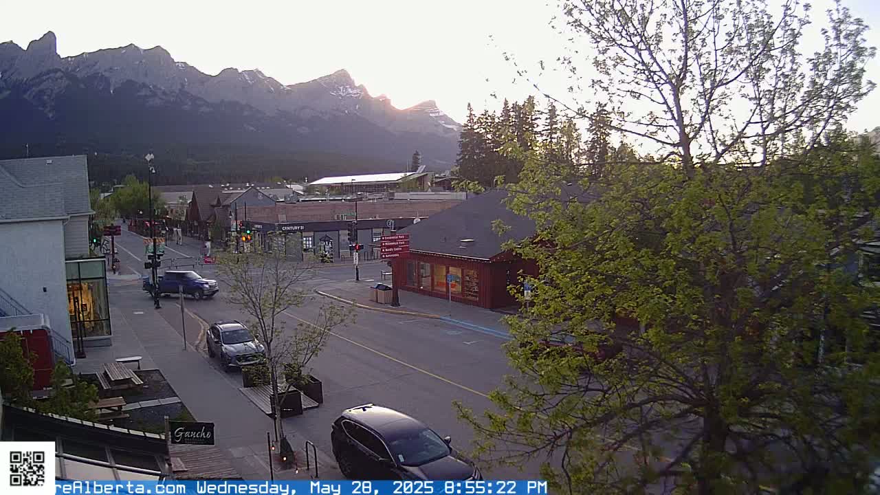 A mountain range is visible in the background of a street scene at sunset, with several cars parked and driving along a road lined with buildings.