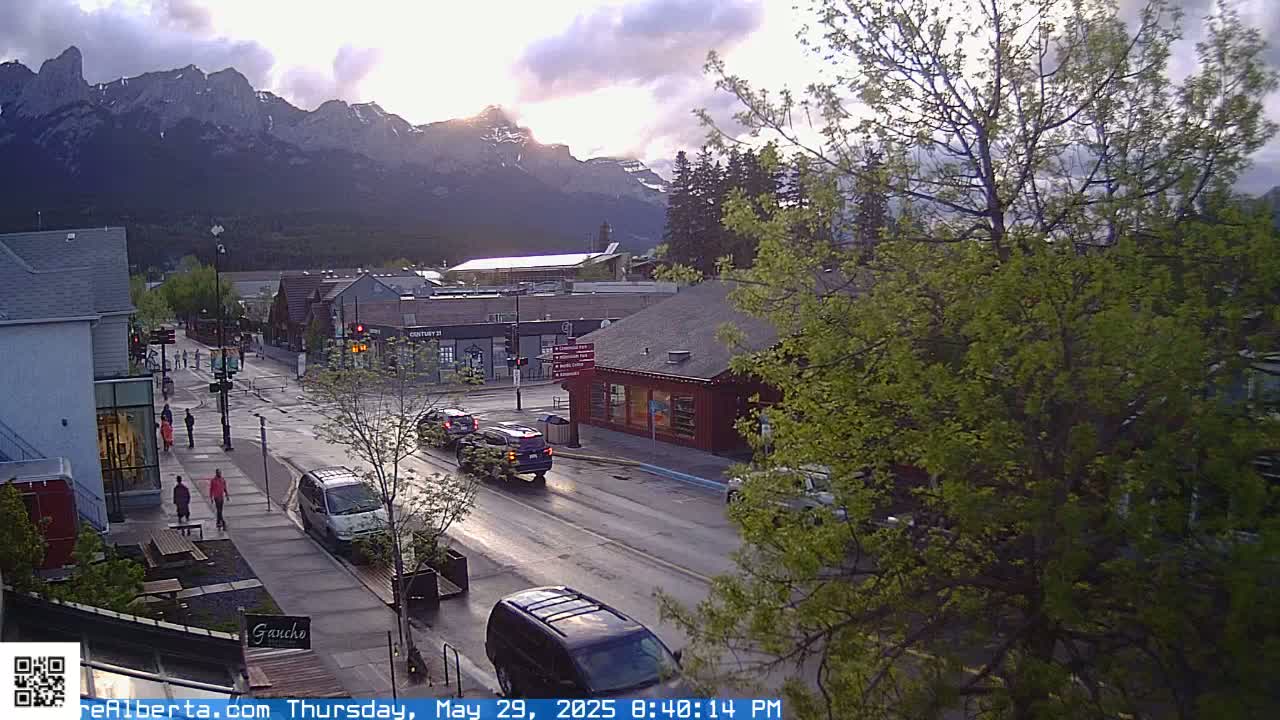 A partly cloudy evening scene shows a town street with cars and pedestrians, overlooked by mountains in the background.