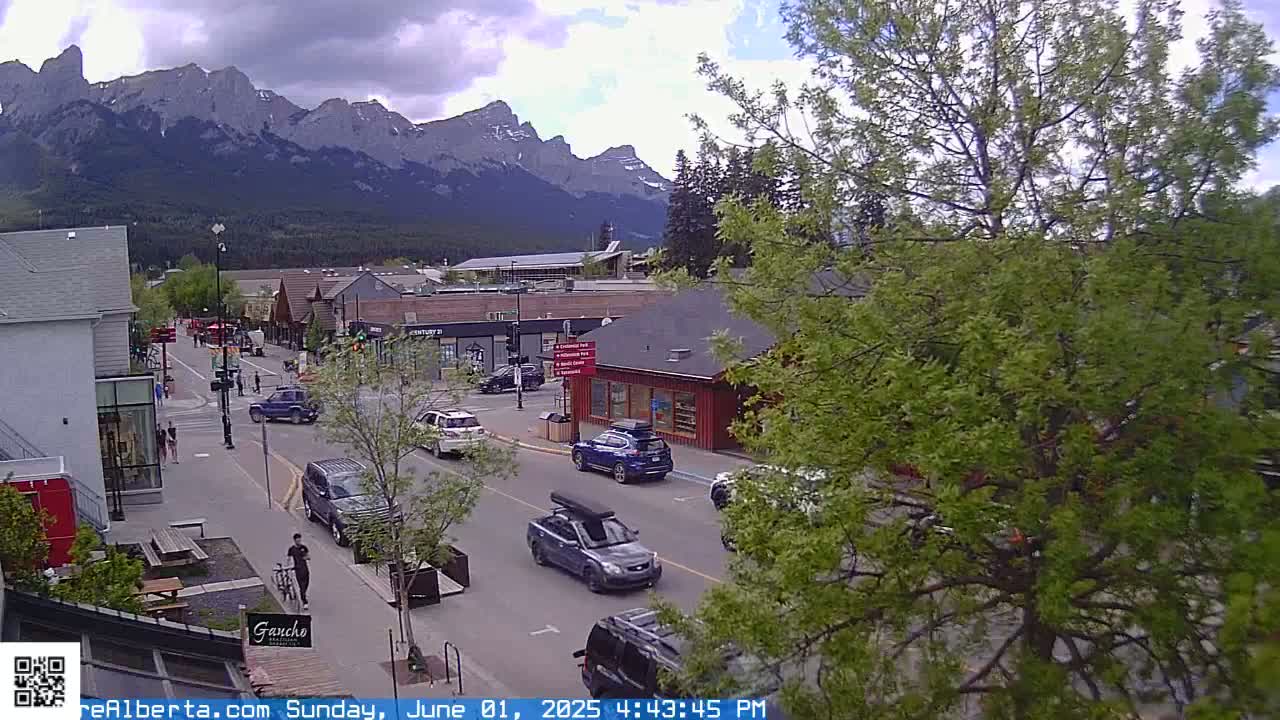 A partly cloudy day reveals a mountain range overlooking a town's street scene with several cars and pedestrians.