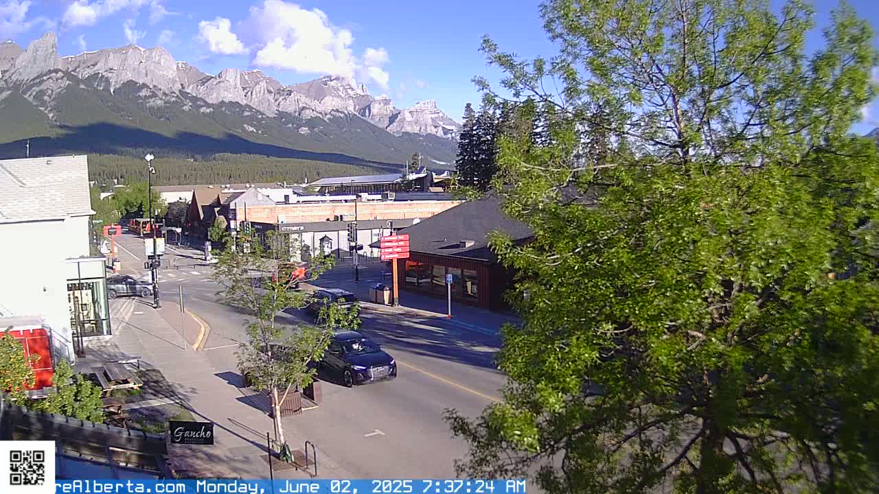 A sunny day reveals a mountain range in the background, overlooking a town's streets with cars driving on them, and lush green trees in the foreground.