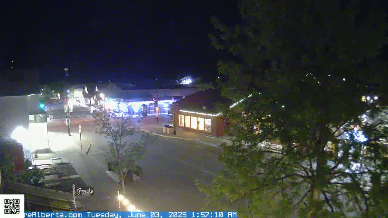 A nighttime, overhead view of a quiet town square with several illuminated buildings and trees.