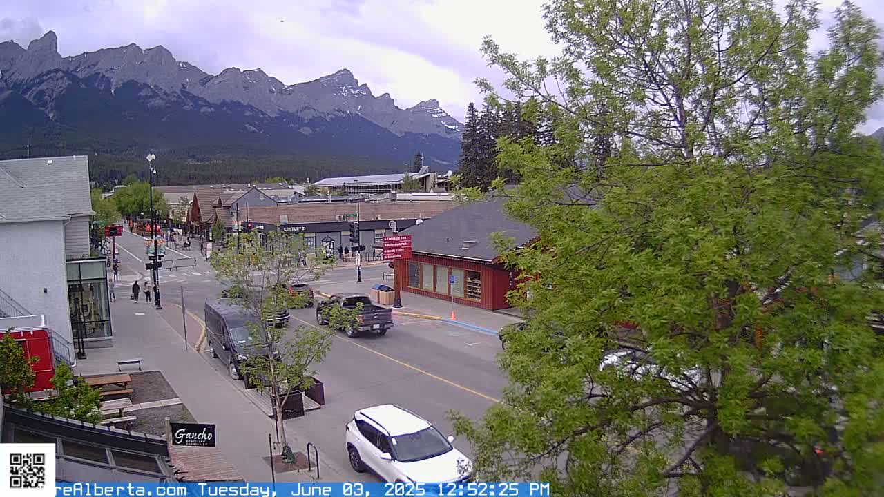 A partly cloudy day reveals a mountain range in the background of a town's street scene, where cars are driving past buildings and pedestrians are walking.