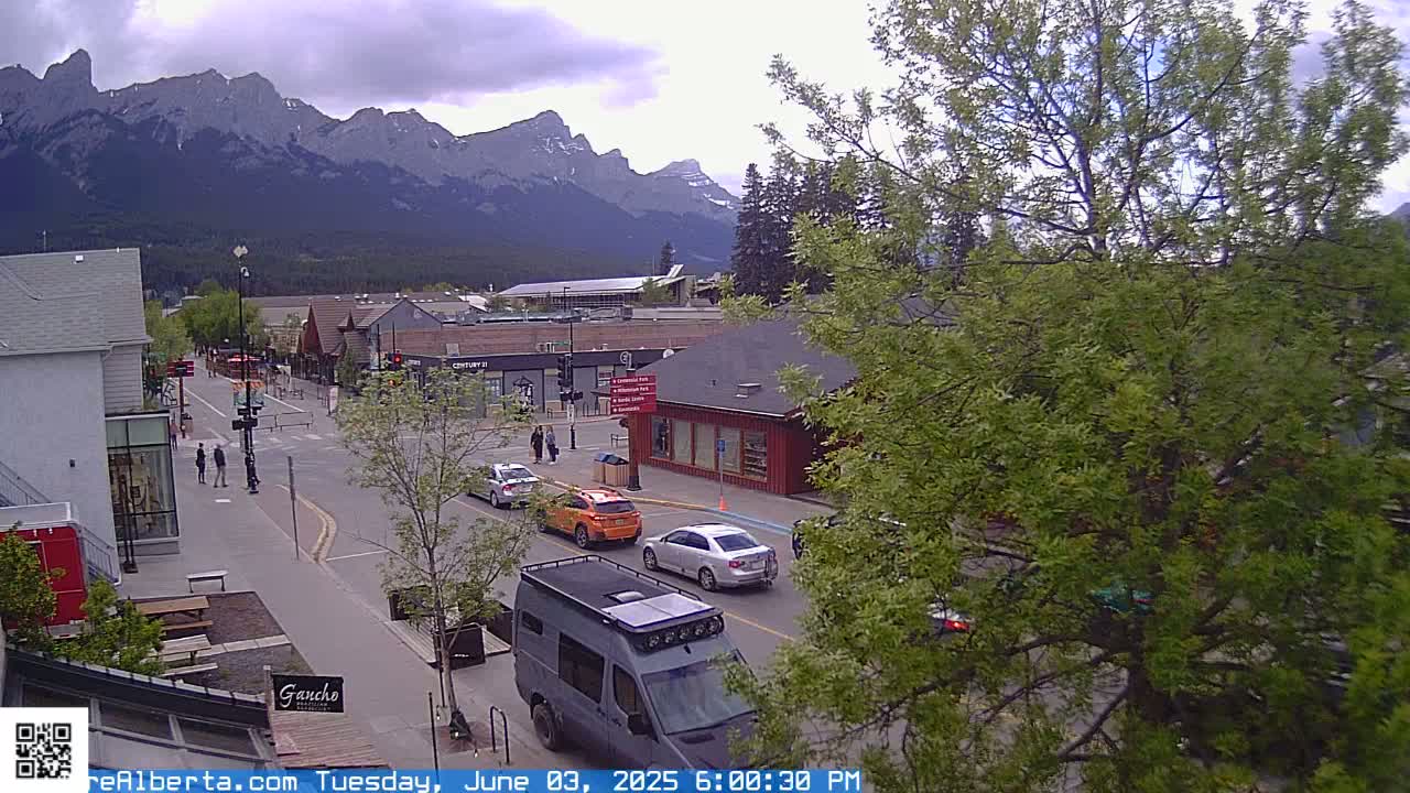 A partly cloudy day reveals a mountain range in the background of a town's street scene where several cars and a van are visible, along with pedestrians on the sidewalk.